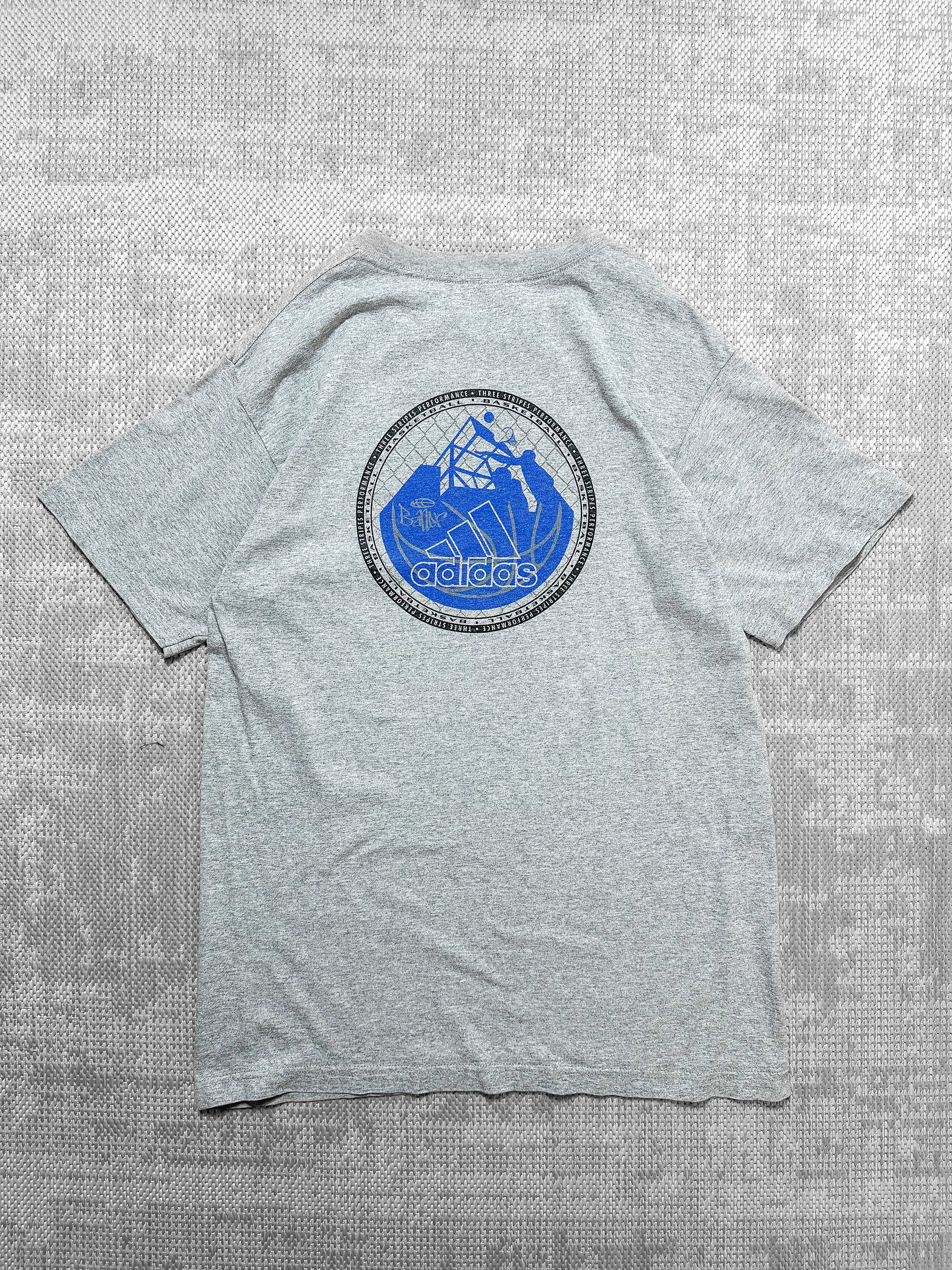 A soft grey short-sleeve T-shirt from Adidas featuring a small logo on the left chest inside a circular badge with the brand's signature text, three stripes, and a blue basketball graphic. The back has a bold circular design with basketball elements like a hoop, player silhouettes, and geometric shapes, framed by the text "Three Stripes Performance Basketball." The T-shirt offers a blend of sporty style and casual wear