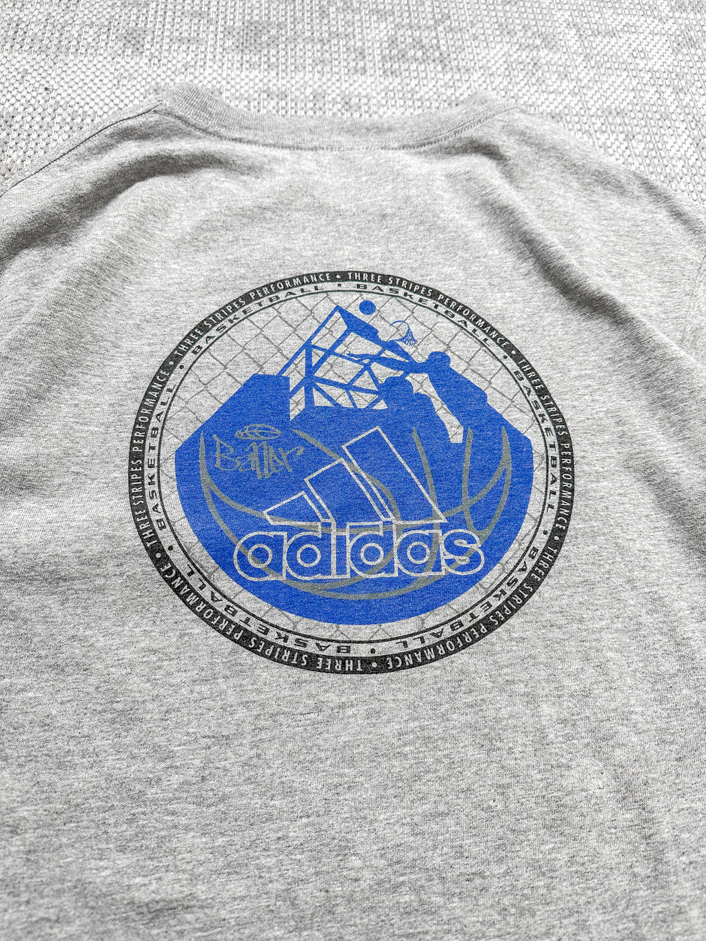 Adidas grey short-sleeve T-shirt with a logo badge on the front, featuring text, stripes, and a blue basketball graphic; bold basketball-inspired design on the back.