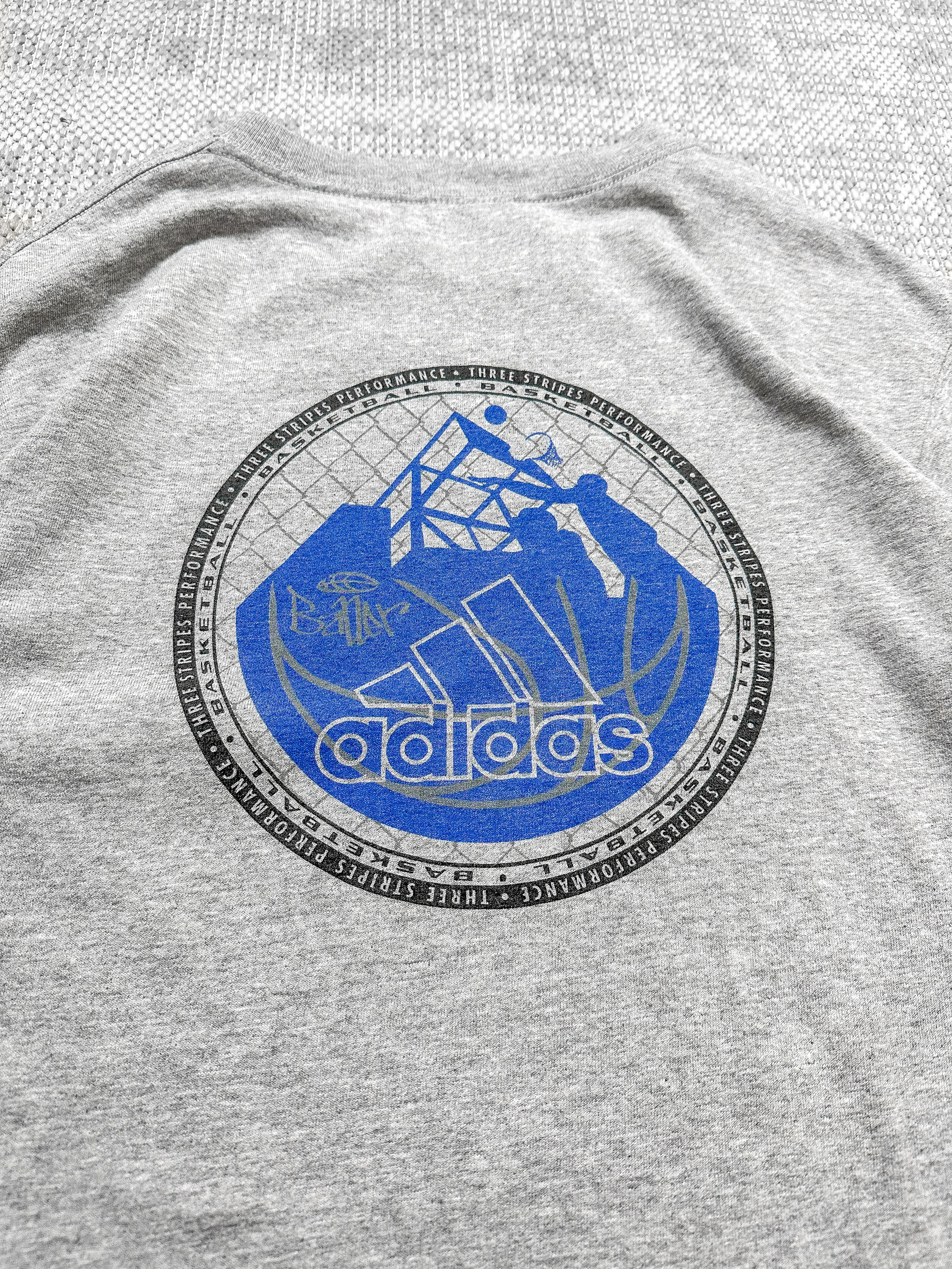Adidas grey short-sleeve T-shirt with a logo badge on the front, featuring text, stripes, and a blue basketball graphic; bold basketball-inspired design on the back.