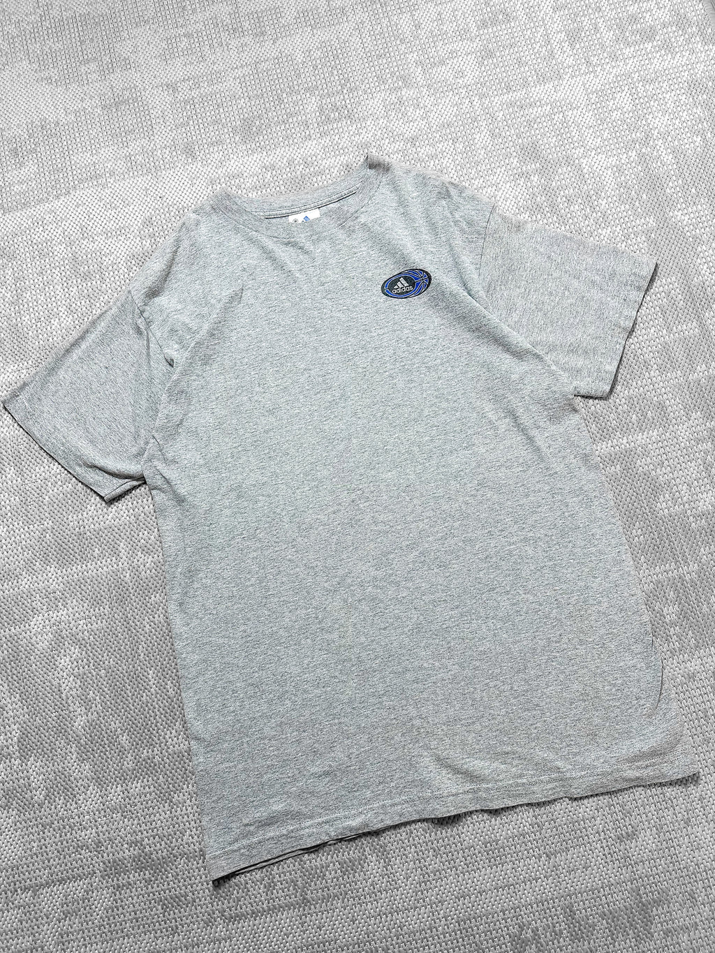 A soft grey short-sleeve T-shirt by Adidas featuring a small logo on the left chest inside a circular badge with the brand's signature text, three stripes, and a blue basketball graphic. The back displays a bold circular graphic with basketball-themed elements, including a hoop, player silhouettes, and geometric patterns, bordered by the text "Three Stripes Performance Basketball." The design combines sporty aesthetics with casual style.