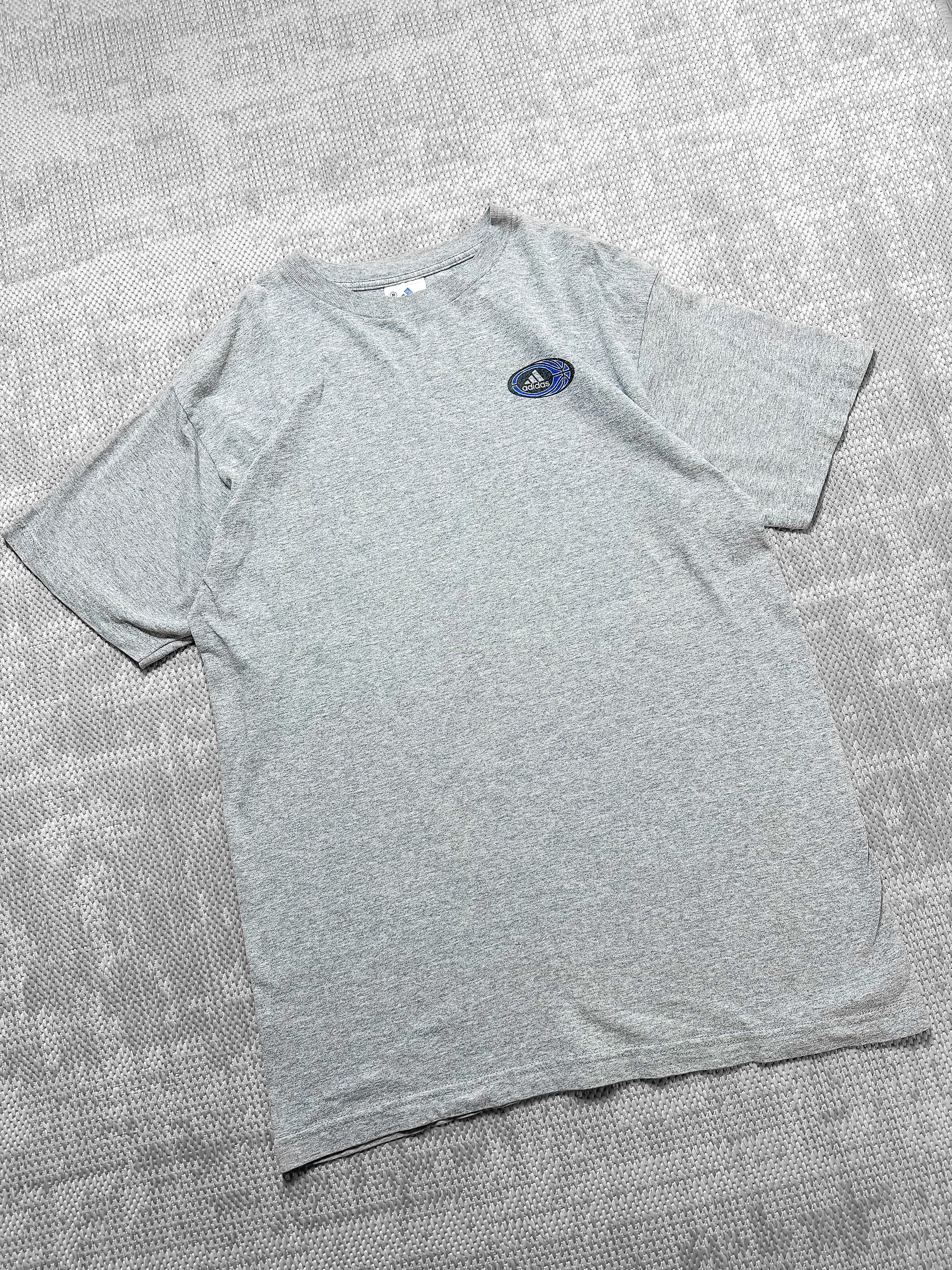A soft grey short-sleeve T-shirt by Adidas featuring a small logo on the left chest inside a circular badge with the brand's signature text, three stripes, and a blue basketball graphic. The back displays a bold circular graphic with basketball-themed elements, including a hoop, player silhouettes, and geometric patterns, bordered by the text "Three Stripes Performance Basketball." The design combines sporty aesthetics with casual style.