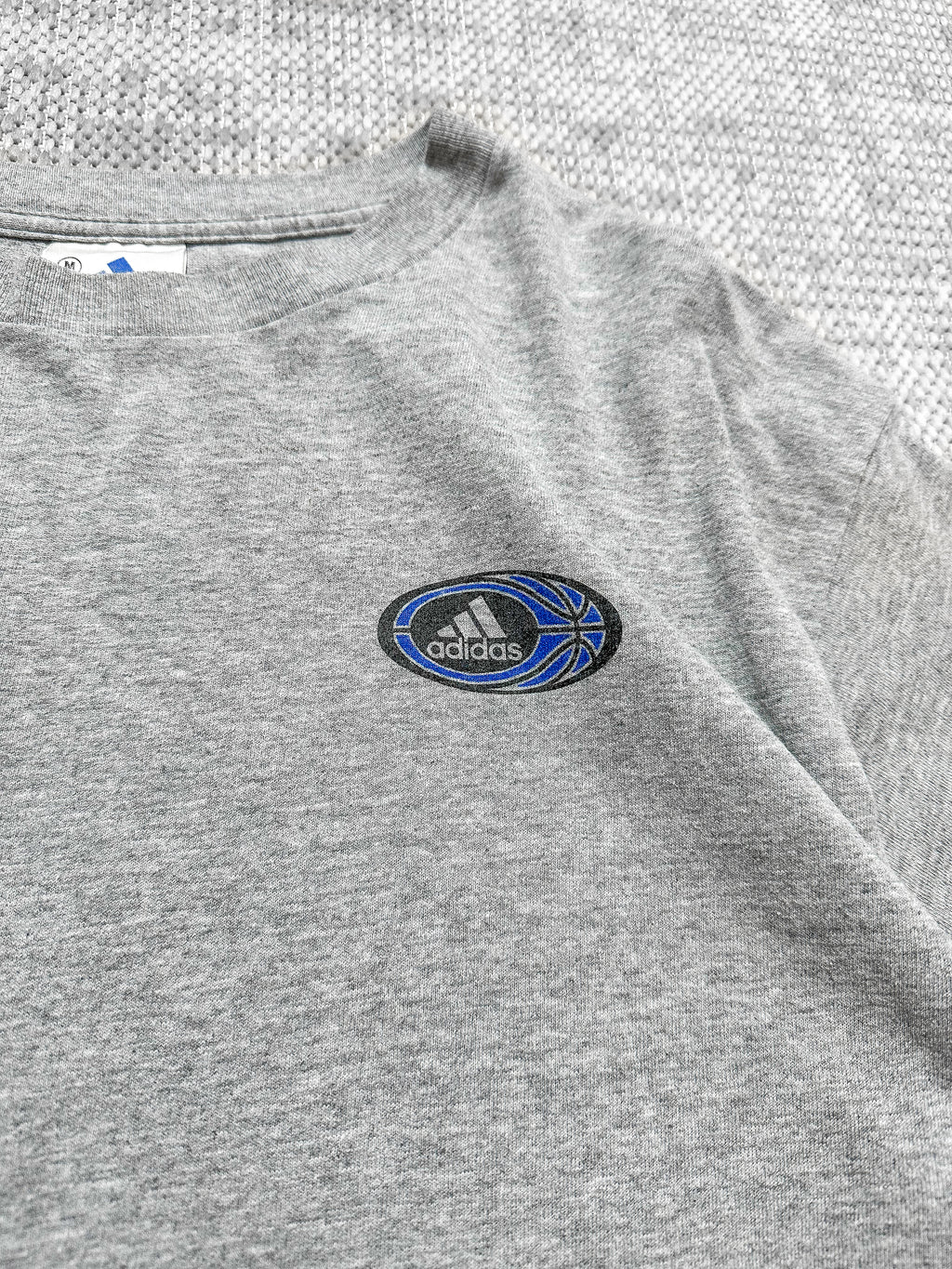 A soft grey short-sleeve T-shirt by Adidas, featuring a small Adidas logo on the left chest inside a circular badge with the brand’s text, three stripes, and a blue basketball graphic. The back displays a bold circular graphic with basketball elements like a hoop, player silhouettes, and geometric patterns, bordered by the text “Three Stripes Performance Basketball.” The T-shirt blends sporty design with casual style.
