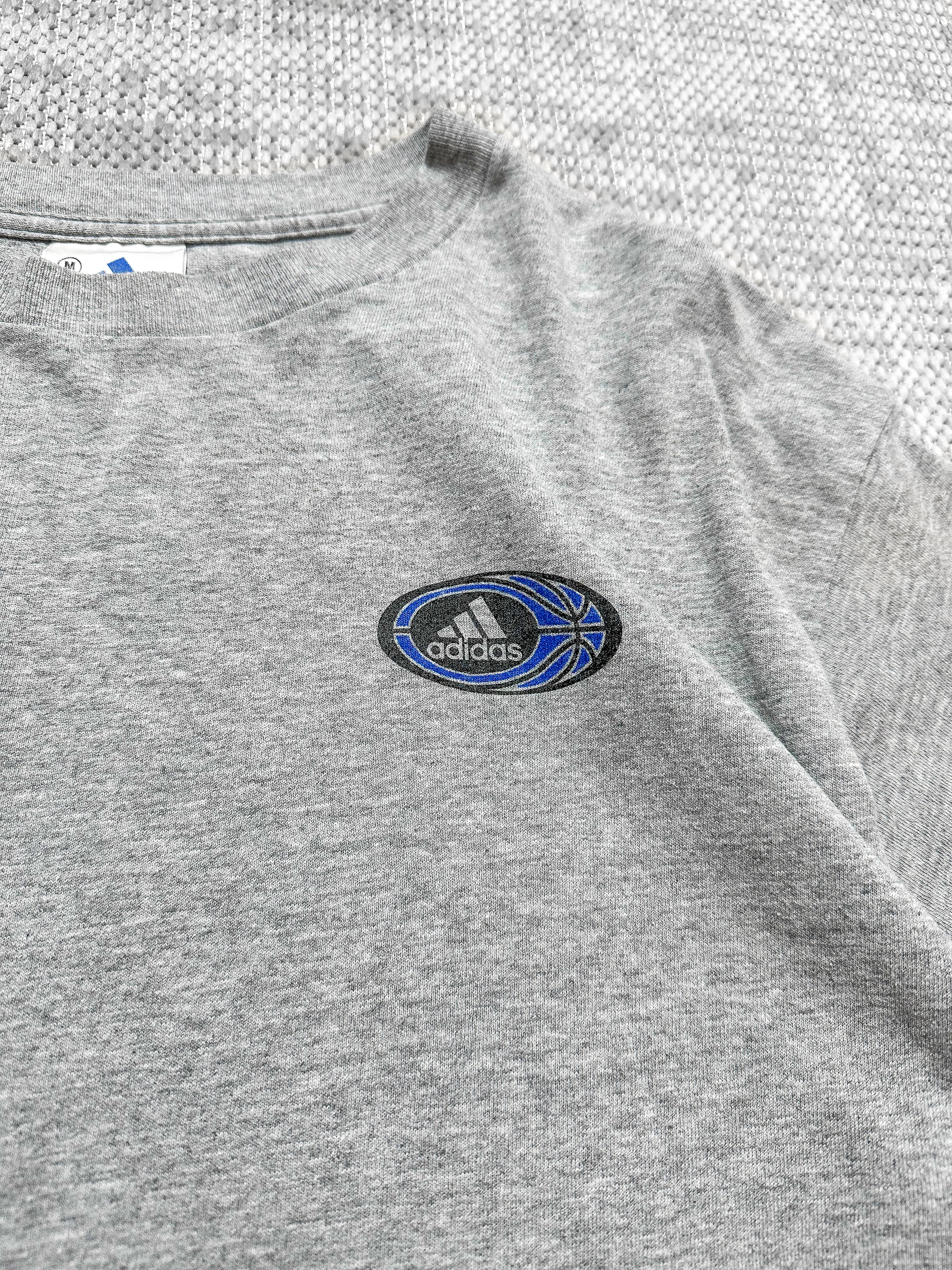 A soft grey short-sleeve T-shirt by Adidas, featuring a small Adidas logo on the left chest inside a circular badge with the brand’s text, three stripes, and a blue basketball graphic. The back displays a bold circular graphic with basketball elements like a hoop, player silhouettes, and geometric patterns, bordered by the text “Three Stripes Performance Basketball.” The T-shirt blends sporty design with casual style.